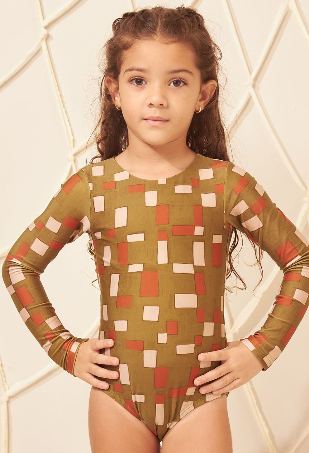 Maiô infantil verde oliva com padrão geométrico bege e laranja, manga longa, modelo mirim de frente com mãos na cintura.