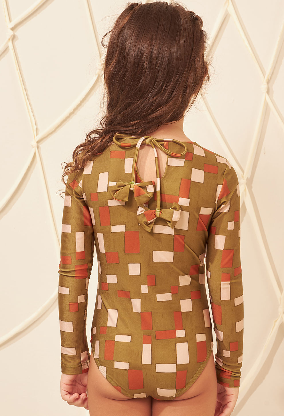 Maiô infantil verde oliva com padrão geométrico vista traseira, detalhes de laços e abertura, modelo mirim com cabelo longo.