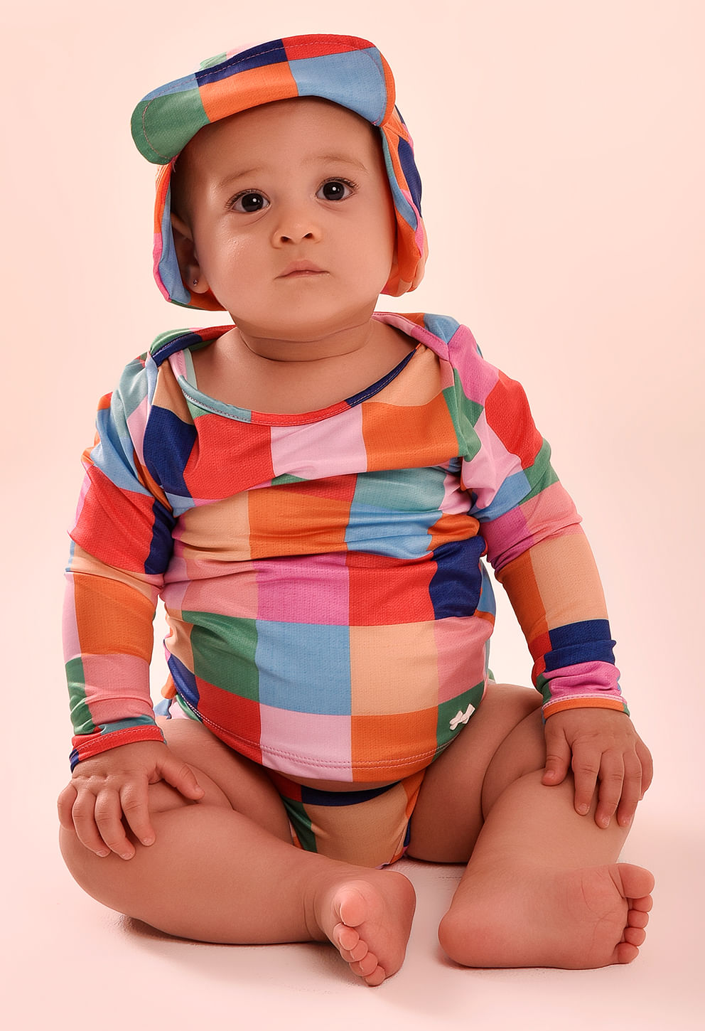 Camisa manga longa bebê com estampa quadriculada colorida e chapéu combinando, bebê sentado em fundo bege claro