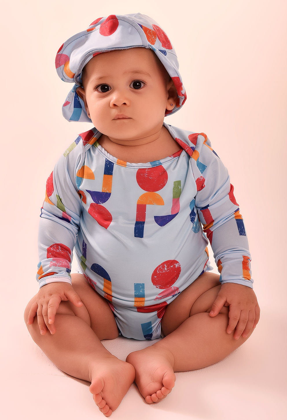 Bebê usando camisa manga longa e chapéu estampados coloridos azul claro.