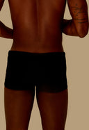 Vista traseira de homem usando sunga short adulto preta com tatuagem no braço em fundo bege.