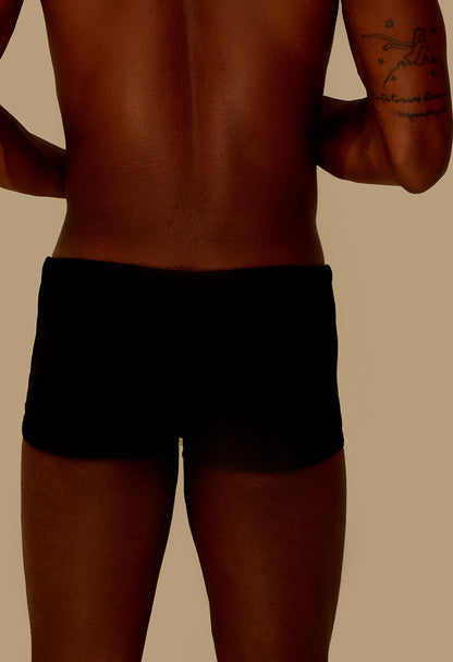 Vista traseira de homem usando sunga short adulto preta com tatuagem no braço em fundo bege.