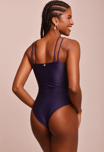 Body feminino azul marinho Yasmim com medalhas douradas nas alças, vista traseira com logo dourado da marca, modelo sorridente em fundo bege.