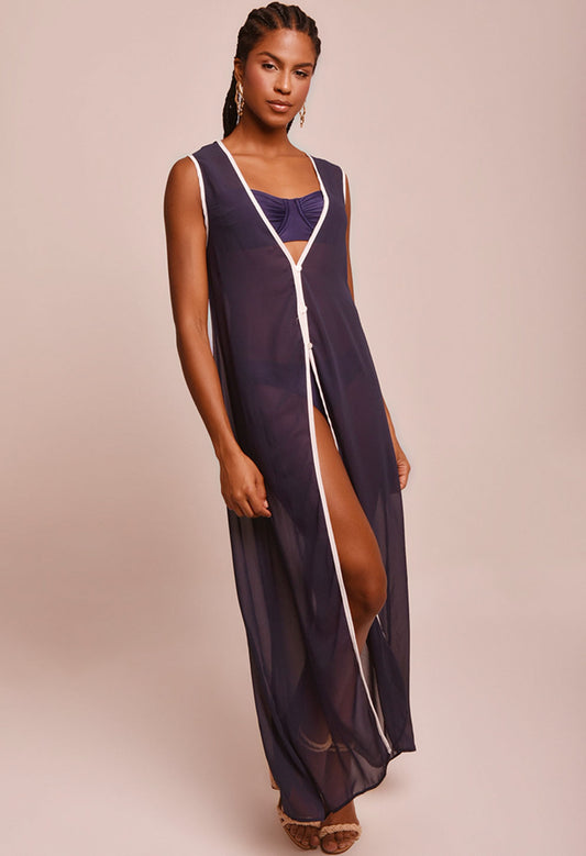 Modelo vestindo saída de praia longa azul marinho transparente com detalhes brancos, com roupa de banho azul escura por baixo, em fundo bege claro.