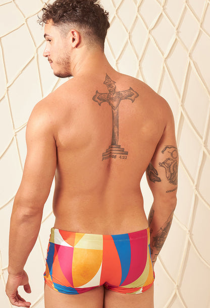 Homem de costas com tatuagens usando sunga short adulto astral colorida, fundo claro e rede decorativa.