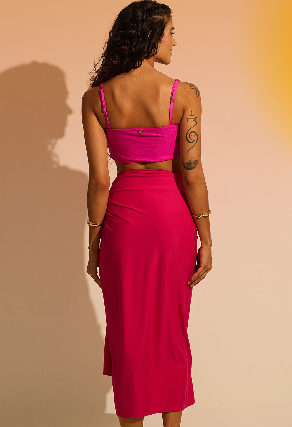 Mulher vista de costas com pareô rosa vibrante longo e top rosa, fundo degradê amarelo e bege.