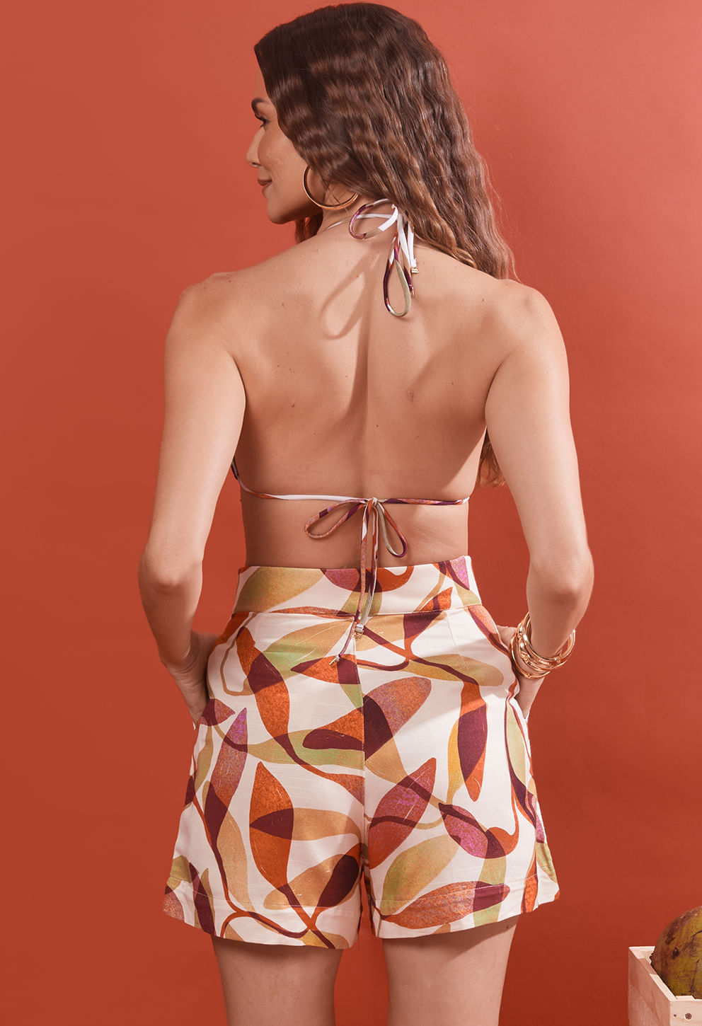 Mulher de costas usando short Milão Cajueiro estampado e top de biquíni, fundo laranja.