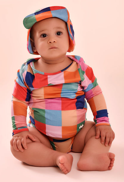 Camisa manga longa bebê com estampa quadriculada colorida e chapéu combinando, bebê sentado em fundo bege claro