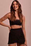 Modelo feminina usando saia mini transpassada preta Saia Mini Transpassada Luar com top faixa preto em fundo neutro.