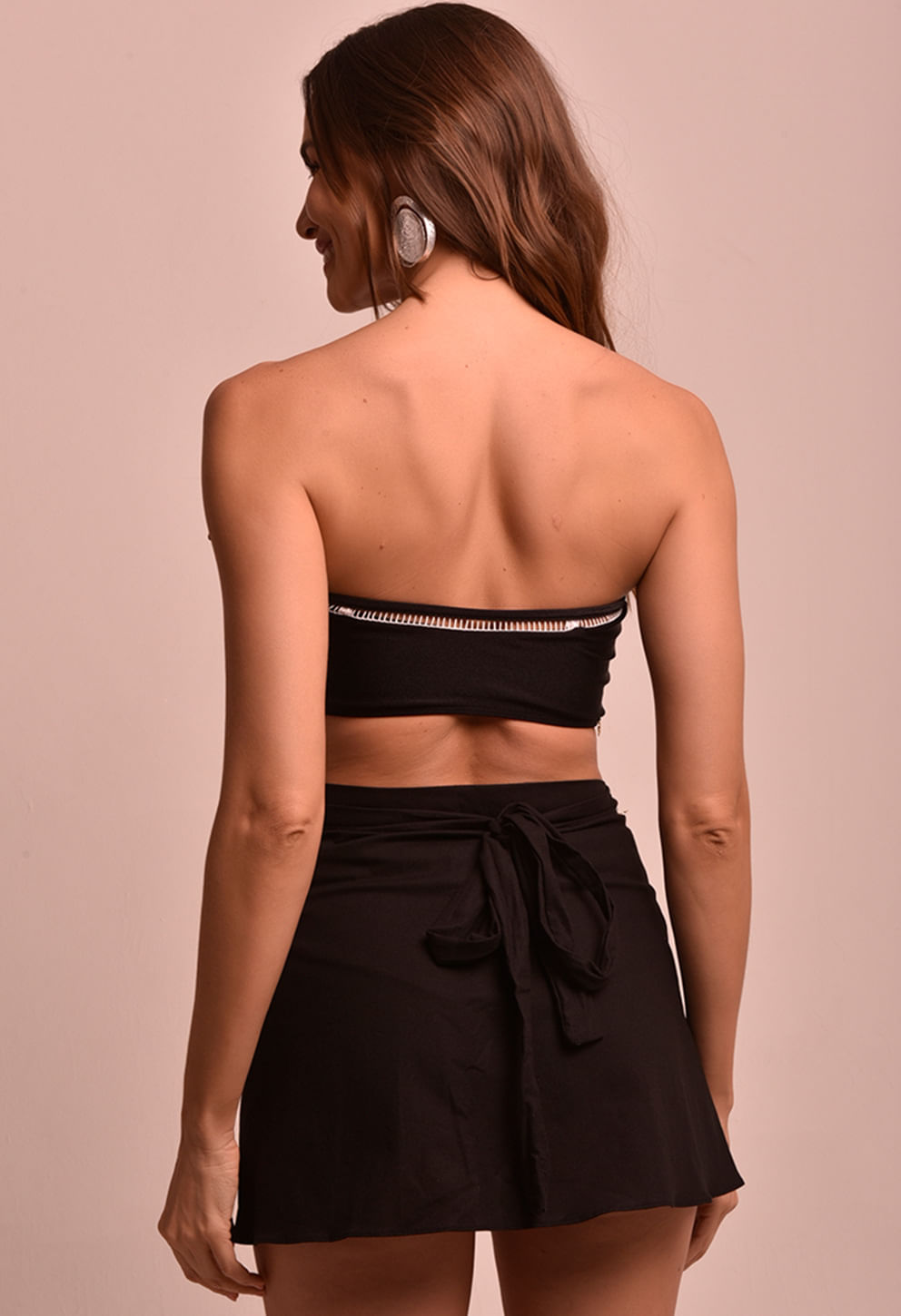 Modelo vista de costas usando saia mini transpassada preta Saia Mini Transpassada Luar com nó na parte de trás e top faixa preto.