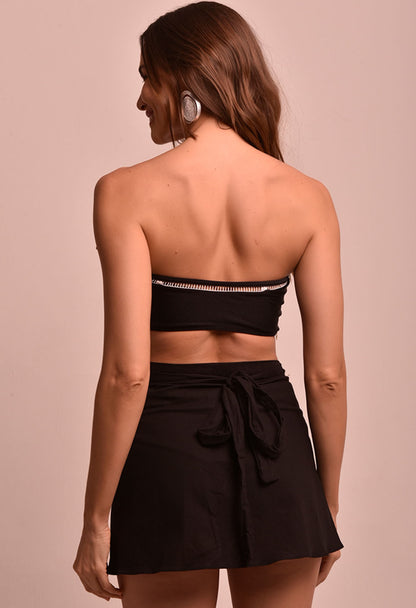 Modelo vista de costas usando saia mini transpassada preta Saia Mini Transpassada Luar com nó na parte de trás e top faixa preto.