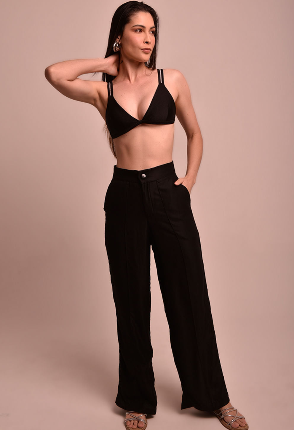 Mulher com calça pantalona preta de cintura alta e top preto, posando para foto em fundo bege, calça da marca Fio a Fio