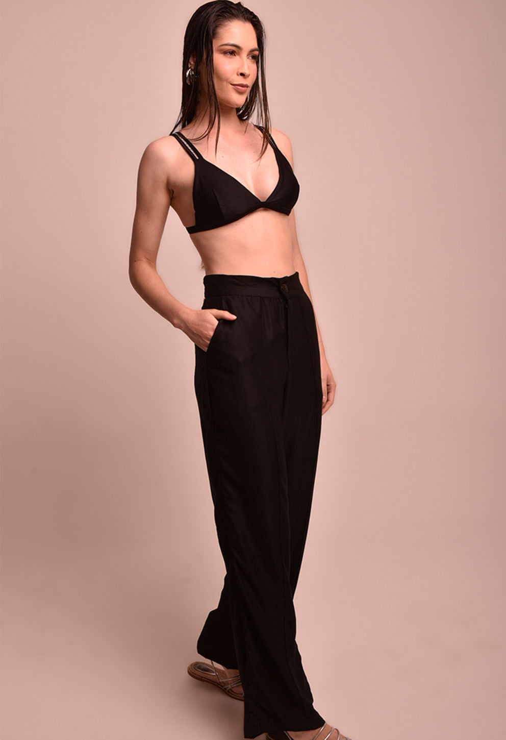 Mulher de lado com calça pantalona preta e top preto, em fundo bege, calça marca Fio a Fio