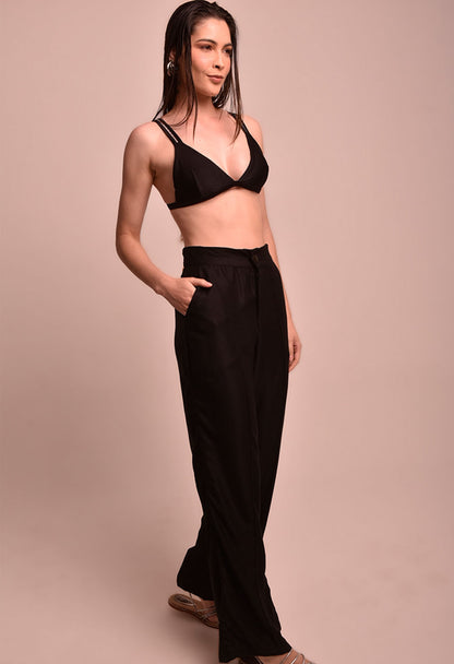 Mulher de lado com calça pantalona preta e top preto, em fundo bege, calça marca Fio a Fio