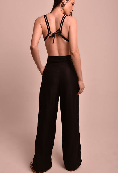 Mulher vista de costas com calça pantalona preta e top preto, posando em fundo bege, calça marca Fio a Fio