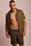 Camisa de linho slim manga curta verde army masculina aberta com bermuda marrom, modelo posando com expressão séria em fundo bege.