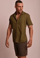 Camisa de linho slim manga curta verde army masculina abotoada com bermuda marrom, modelo olhando para esquerda em fundo bege.