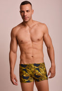 Modelo masculino vestindo sunga short adulto com estampa de folhas amarelas e verdes sobre fundo preto, fundo bege neutro.