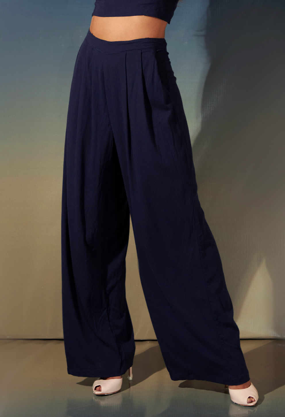 Calça pantalona básica marinho com pregas frontais em destaque com sapato de salto branco, fundo degradê neutro.