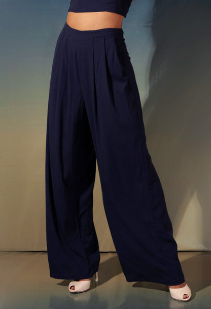 Calça pantalona básica marinho com pregas frontais em destaque com sapato de salto branco, fundo degradê neutro.