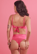 Vista traseira de mulher com biquíni rosa pink acetinado com amarração nas costas e calcinha hot pants em fundo rosa.