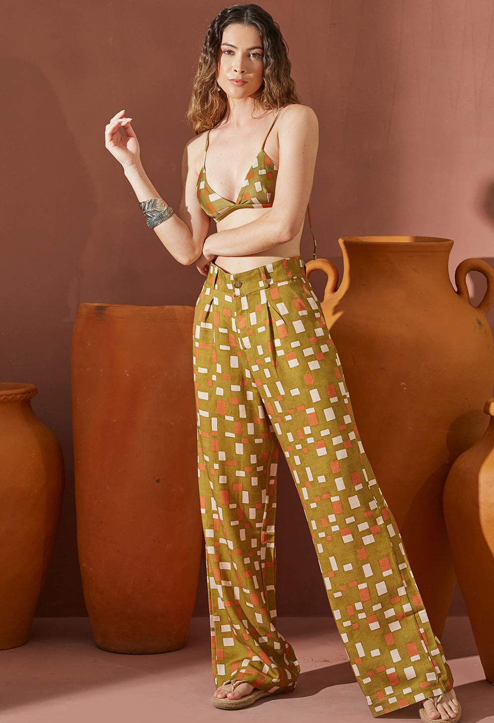 Mulher vestindo pantalona social Ilha estampada em verde oliva com padrão geométrico, combinada com top de alças finas, posando em fundo com vasos de cerâmica.