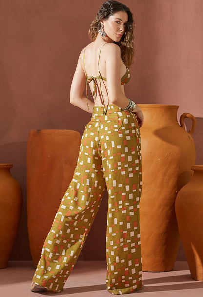 Mulher de costas vestindo pantalona social Ilha estampada verde oliva com padrão geométrico, combinada com top amarrado, posando em fundo com vasos de cerâmica laranja.